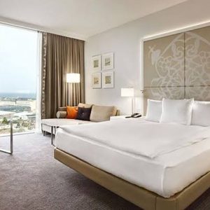 A modern, elegantly decorated hotel room with a large bed, a sofa, a floor lamp, and a large window offering a city view.
