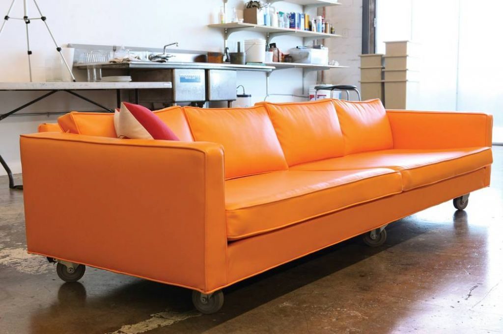 Sofá moderno de color naranja brillante con ruedas, situado en un estudio con estanterías y equipo en el fondo.